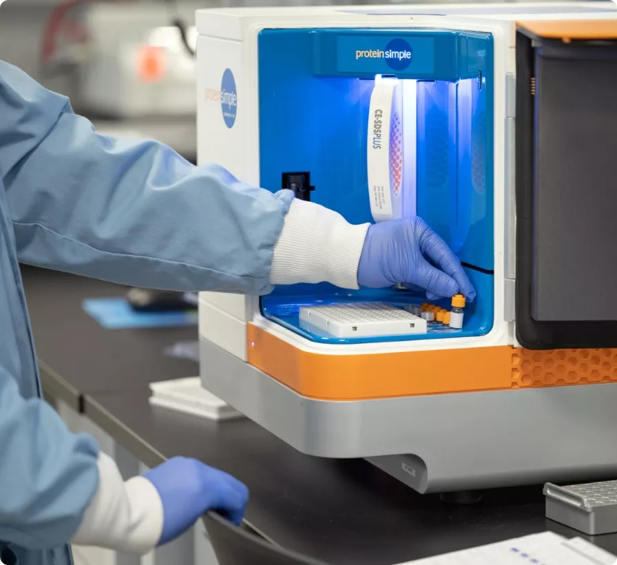 A scientist in a lab coat and gloves loads samples into a ProteinSimple instrument for analysis.